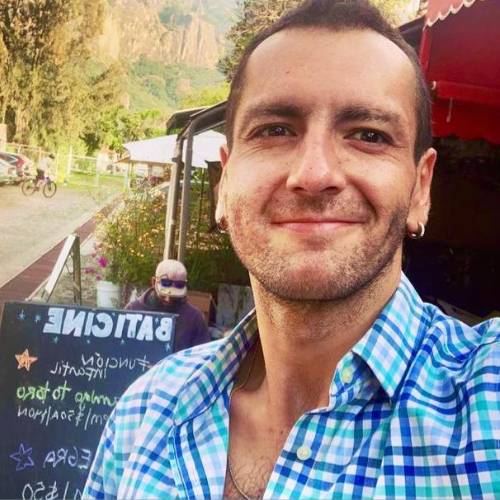 Hombre sonriendo en un entorno natural, con una camisa de cuadros, frente a un cartel de "Batience" y una persona al fondo, reflejando un estilo de vida saludable y conexión con la naturaleza.